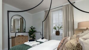 Un dormitorio con una cama con dosel, un aparador de madera, un espejo, una ventana con cortinas y una lámpara de noche.