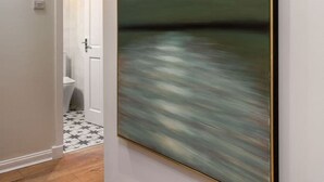 Un pasillo con un cuadro abstracto grande en la pared, suelo de madera y una puerta que da a un baño con suelo de baldosas estampadas.