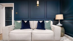 A beige sofa with dark and light pillows, a floor lamp, and a dark blue wall with paneling. - Sapphire Suite The Stirling Residence (Stirling)