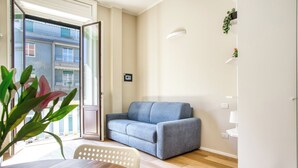 A modern living room with a grey sofa, a wooden dining table, and a white chair. There is a door leading to a balcony with a view of buildings. - JOIVY Cute Central Studio (Milan)