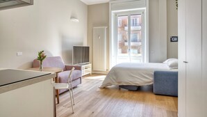 A modern hotel room with a bed, a desk, a chair, a TV, and a view of the city through a large window. - JOIVY Cute Central Studio (Milan)