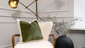 A modern chair with a white, textured seat and green and white cushions, positioned next to a black vase with branches, under a black floor lamp with three light fixtures. - Emerald Suite, The Stirling Residence (Stirling)