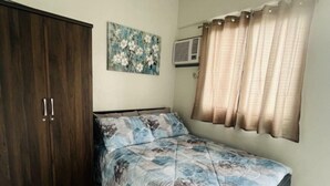 A bedroom with a bed, a wardrobe, a painting, a window with curtains, and an air conditioning unit. - Charming 1 bedroom apartment with AC, WiFi in lovely Lapu-Lapu City (Lapu-Lapu City)