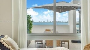 A room with a large window overlooking a cityscape and water. There is a bed with a white headboard, a small table, and a chair. - Luxury Penthouse with Water Views and  Massive Private Terrace and Free Parking (Miami)