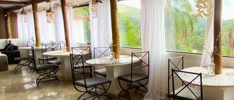A restaurant with round tables and chairs, large windows with curtains, and a tiled floor.