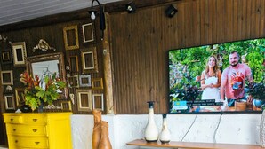 A living room with a TV displaying a couple, a yellow dresser, and a wooden wall panel. - Pousada Encantos da Serra Macacos (Nova Lima)