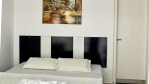 A neatly made bed with a white bedspread and pillows, a painting of a landscape above it, and a closed door to the right. - Pousada Encantos da Serra Macacos (Nova Lima)