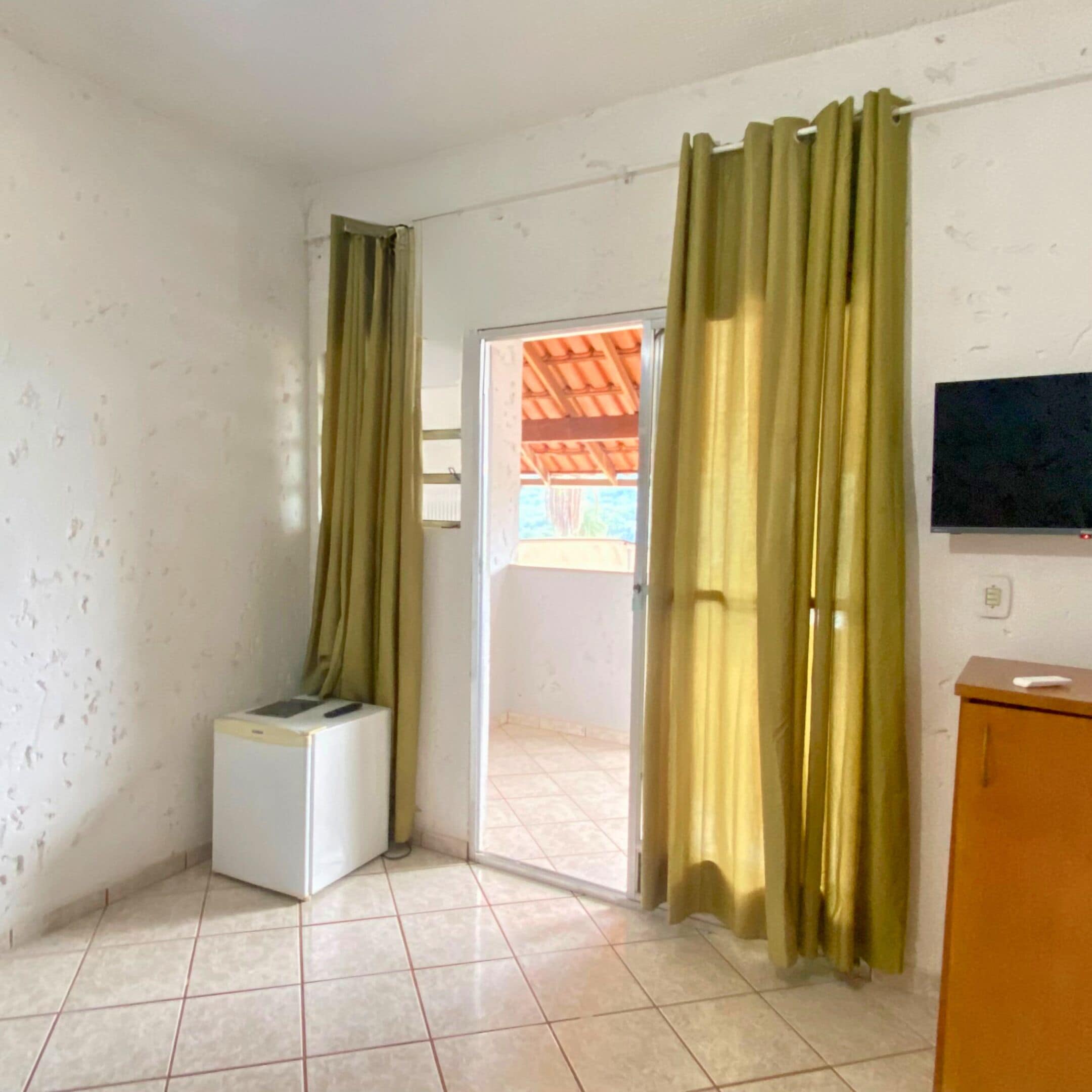 Une pièce dotée d’un sol carrelé, d’une petite armoire blanche, d’un téléviseur fixé au mur et d’une porte-fenêtre coulissante donnant sur un espace extérieur.