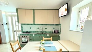 Een kleine keuken met een eettafel, stoelen, een aan de muur bevestigde televisie en een raam met bloemen.
