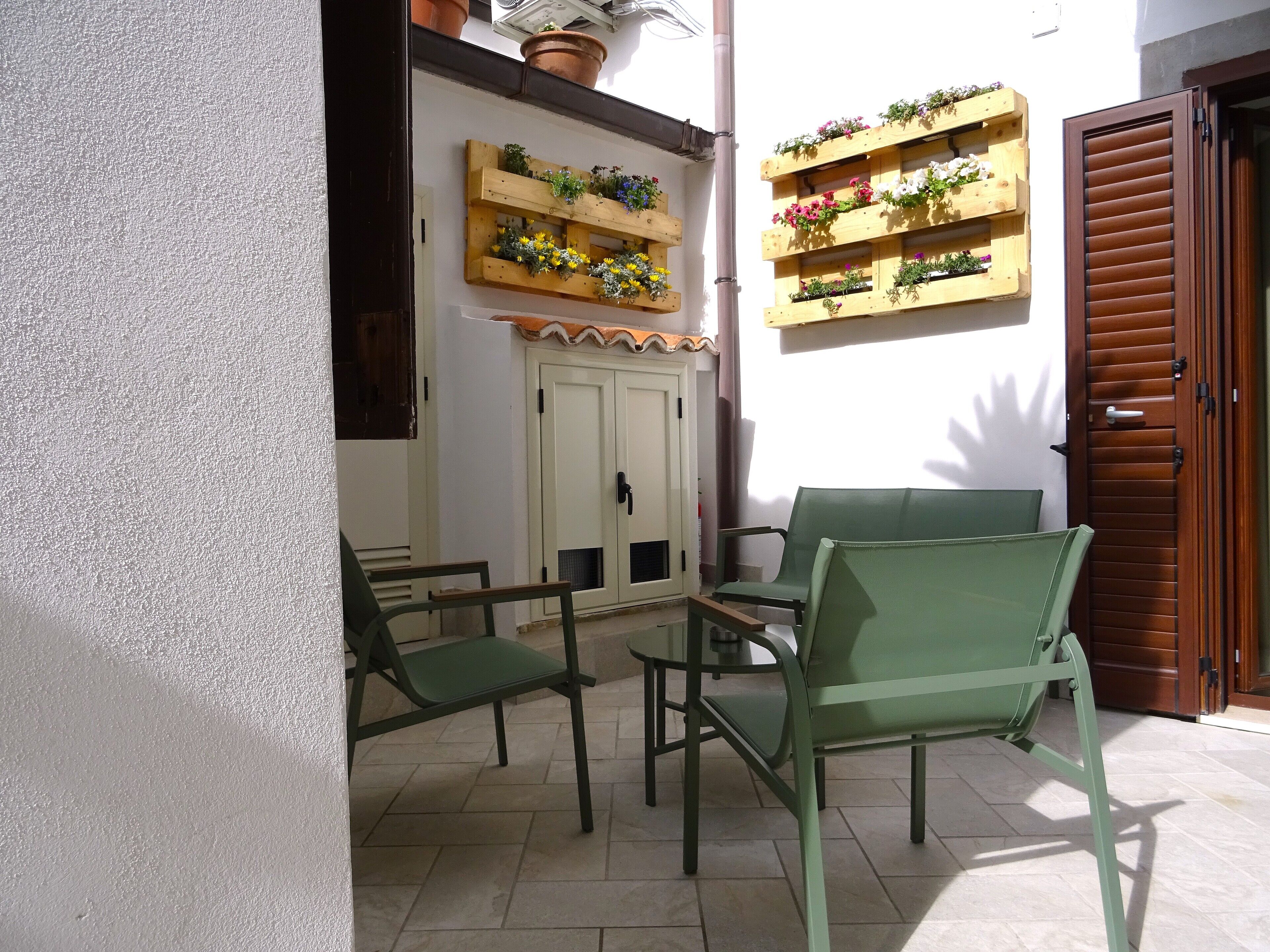 Un espace patio avec des chaises vertes et une table, des jardinières en bois ornées de fleurs fixées au mur, et une porte en bois.