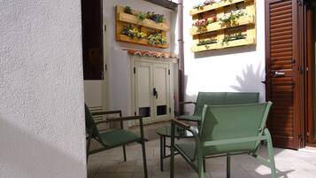 Un espace patio avec des chaises vertes et une table, des jardinières en bois ornées de fleurs fixées au mur, et une porte en bois.