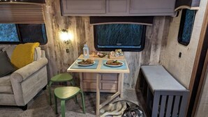 A cozy RV interior with a dining area, a sofa, a dog, and a window with a view of trees.