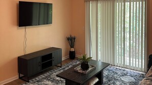 Un salon doté d’un ventilateur de plafond, d’un téléviseur à écran plat fixé au mur, d’un meuble de rangement en bois foncé, d’un tapis à motifs, d’une table basse agrémentée d’objets décoratifs et d’une grande fenêtre avec des stores verticaux.