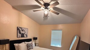 Une chambre à coucher comprenant un lit, un ventilateur de plafond, une table de chevet, une lampe, un téléviseur et une fenêtre avec des stores.