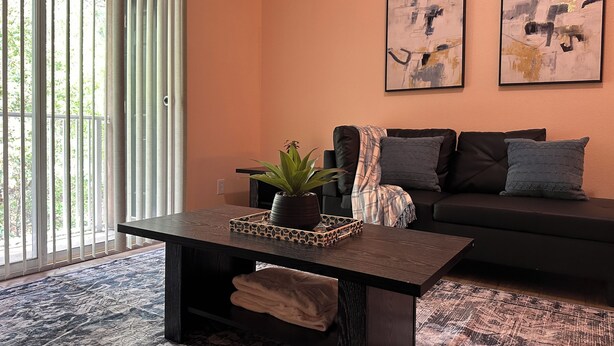 Un salon moderne avec une table basse en bois foncé, une plante en pot et un canapé noir agrémenté de coussins gris. Les murs sont ornés de tableaux abstraits et une grande fenêtre est dotée de stores.
