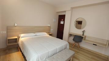 A modern bedroom with a large bed, bedside tables, a bench, a desk with a chair, a mirror, and a door with a sign.