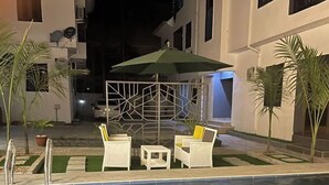 Ein Poolbereich mit zwei weißen Liegestühlen und einem kleinen Tisch, umgeben von Palmen und einem geometrischen Zaun.