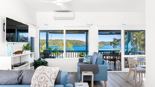 A modern living room with a large TV, comfortable seating, and a view of a lake and hills.