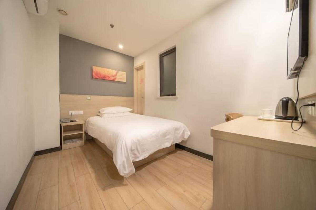 A hotel room with a bed, bedside table, a small desk with a chair, a coffee machine, and a painting on the wall.