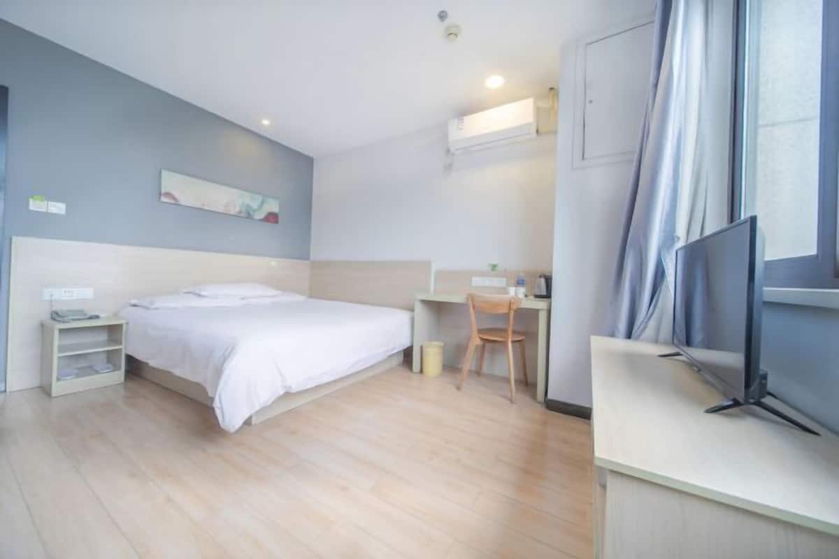A modern hotel room with a bed, desk, chair, TV, and a painting on the wall.