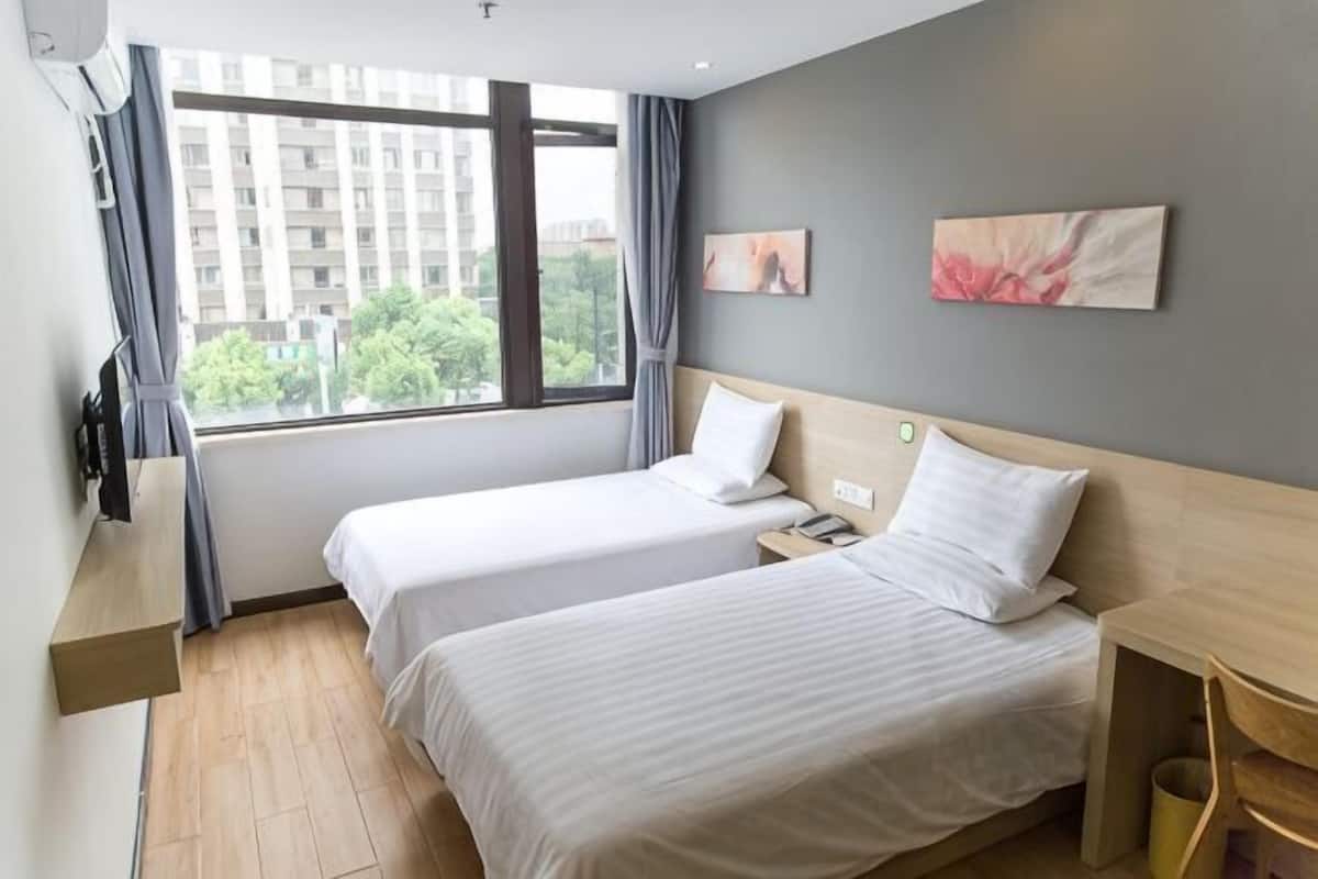 A hotel room with two beds, a TV, a desk, and a window with a city view.