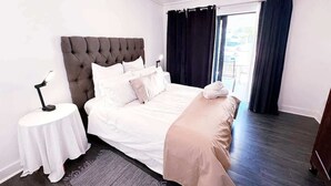 A bedroom with a tufted headboard, a round bedside table, a bed with white bedding and a beige throw, and a view of the outdoors through a window with dark curtains. - Spacious 1 Bedroom Apartment, Atlantica, Seapoint (Cape Town)