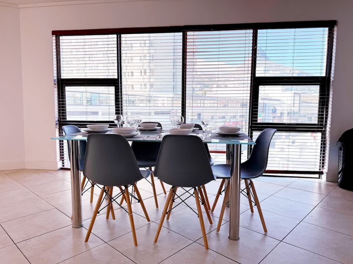 A dining area with a table set for four, large windows with blinds, and a tiled floor.