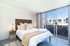 Ein modernes Schlafzimmer mit einem großen Bett, einem Holzkopfstück und Blick auf Gebäude durch große Fenster.