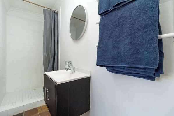 A bathroom with a shower, a sink, a mirror, and a stack of towels.