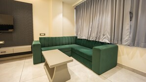 A modern living room with a green sectional sofa, a small coffee table, a flat-screen TV mounted on a dark wall, and a window with grey curtains. - Hotel Shankar Palace Beawar (Beawar)