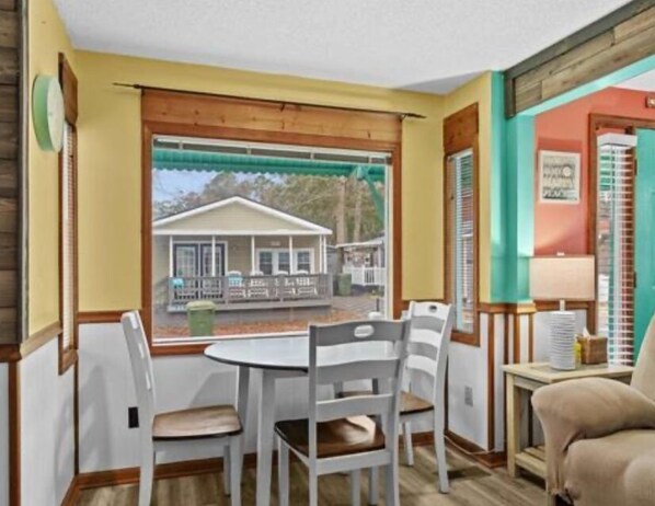 A room with a wooden-framed window, a small table with chairs, and a view of a house outside. - Beckham by The Sea (Myrtle Beach)