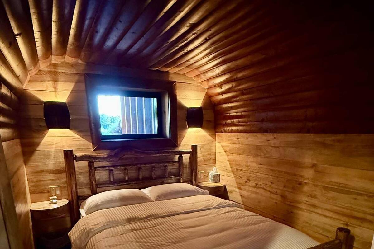 Ein gemütliches Schlafzimmer in einer Holzhütte mit einem Bett, zwei Nachttischen, einem Fenster und einer Wandlampe.