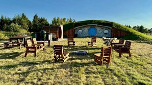 Un área de estar al aire libre de estilo Hobbit con bancos y mesas de madera, un muro de piedra con una puerta azul y un campo cubierto de césped.