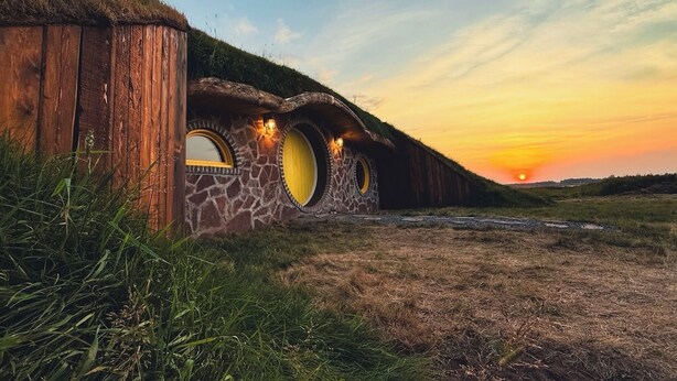 Una casa de estilo Hobbit con puertas redondas y techo de paja, ubicada contra un atardecer.