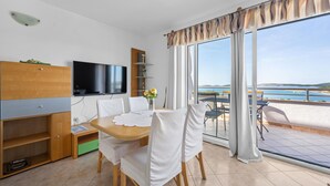 Una sala moderna con televisor de pantalla plana, muebles de madera, una mesa de comedor con sillas blancas y un balcón con vista al mar.