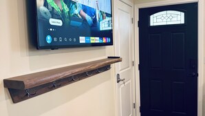 Un téléviseur à écran plat fixé au mur, au-dessus d’une étagère en bois munie de crochets, une porte avec une fenêtre et un paillasson de couleur sombre à l’entrée.