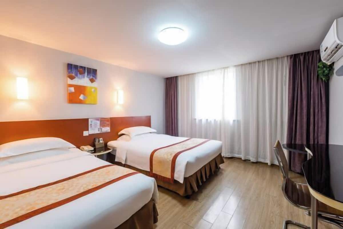 Ein Hotelzimmer mit zwei Betten, einem Schreibtisch und einem Stuhl. An der Wand hängt ein Bild und ein Fenster mit Vorhängen.