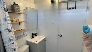 Casa de banho com sanita, lavatório e chuveiro. Existem prateleiras com objetos decorativos e um porta-toalhas.