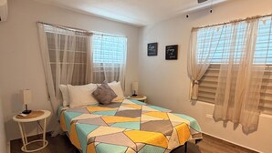 Un dormitorio con cama, ventilador de techo, mesita de noche y ventana con cortinas de encaje.