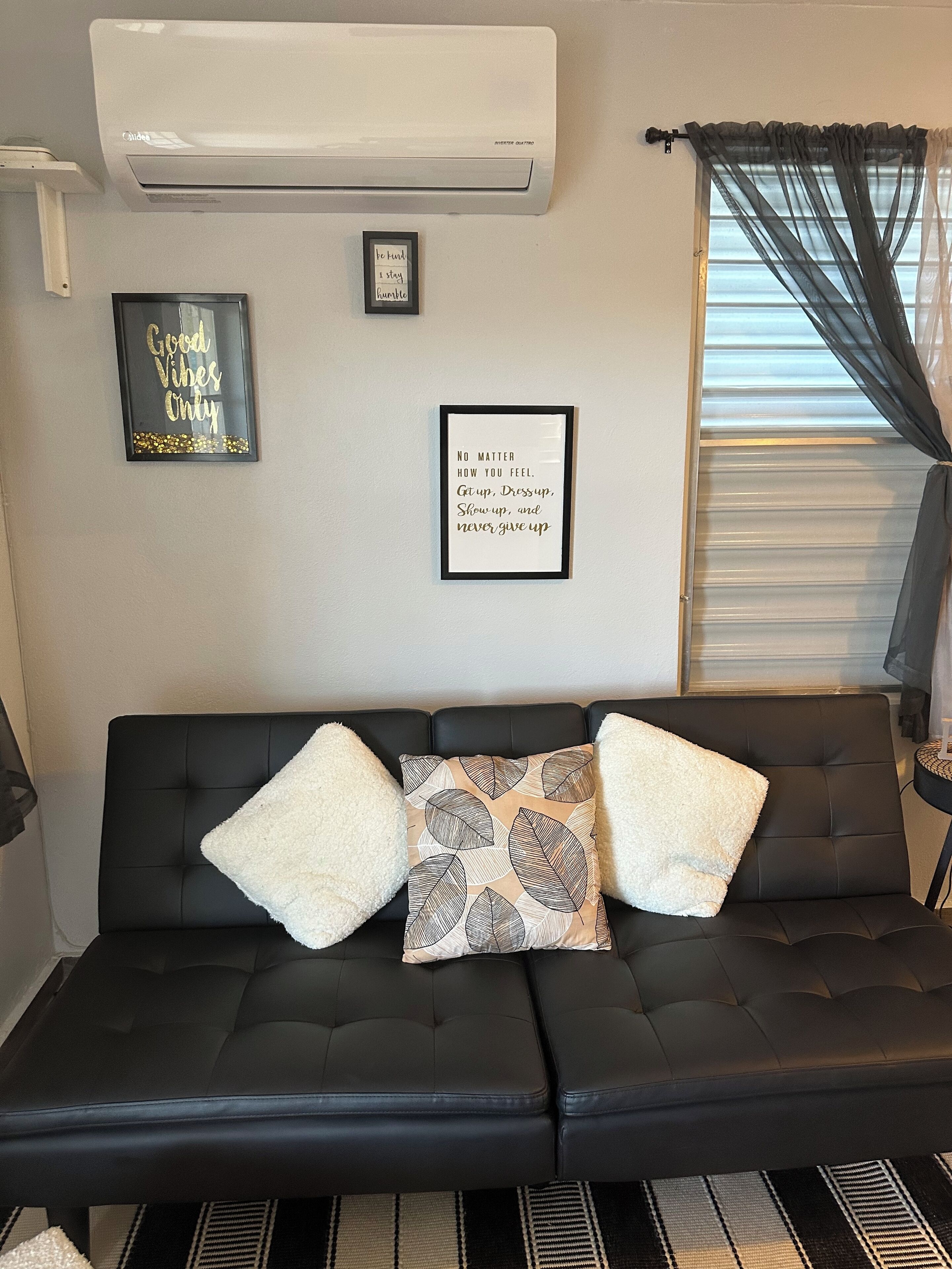 A black leather sofa with three pillows, two white fluffy and one patterned. There are two framed motivational quotes on the wall.