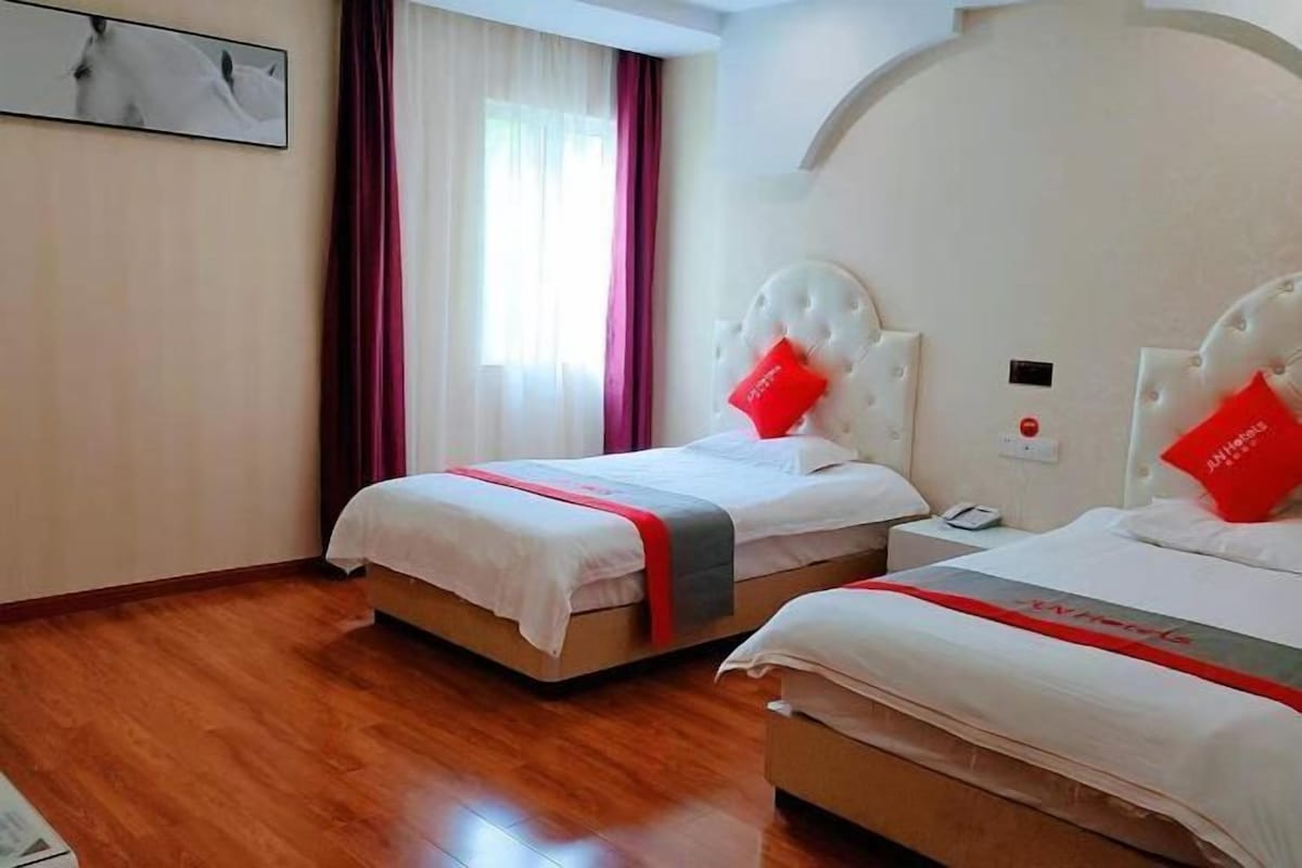 A hotel room with two beds, a wooden floor, a white headboard, and a red accent pillow.