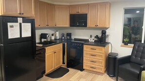 A compact kitchen with wooden cabinets, a black refrigerator, a stove, and a microwave. There is a black chair and a small trash bin. - Wisc Dells Romantic Getaway for two (Baraboo)