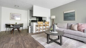 A modern living room with a grey sofa, a dining table with chairs, a small coffee table, and a kitchen area with white cabinets and a black countertop. - Landing Winston-Salem near Wake Forest University (Winston-Salem)