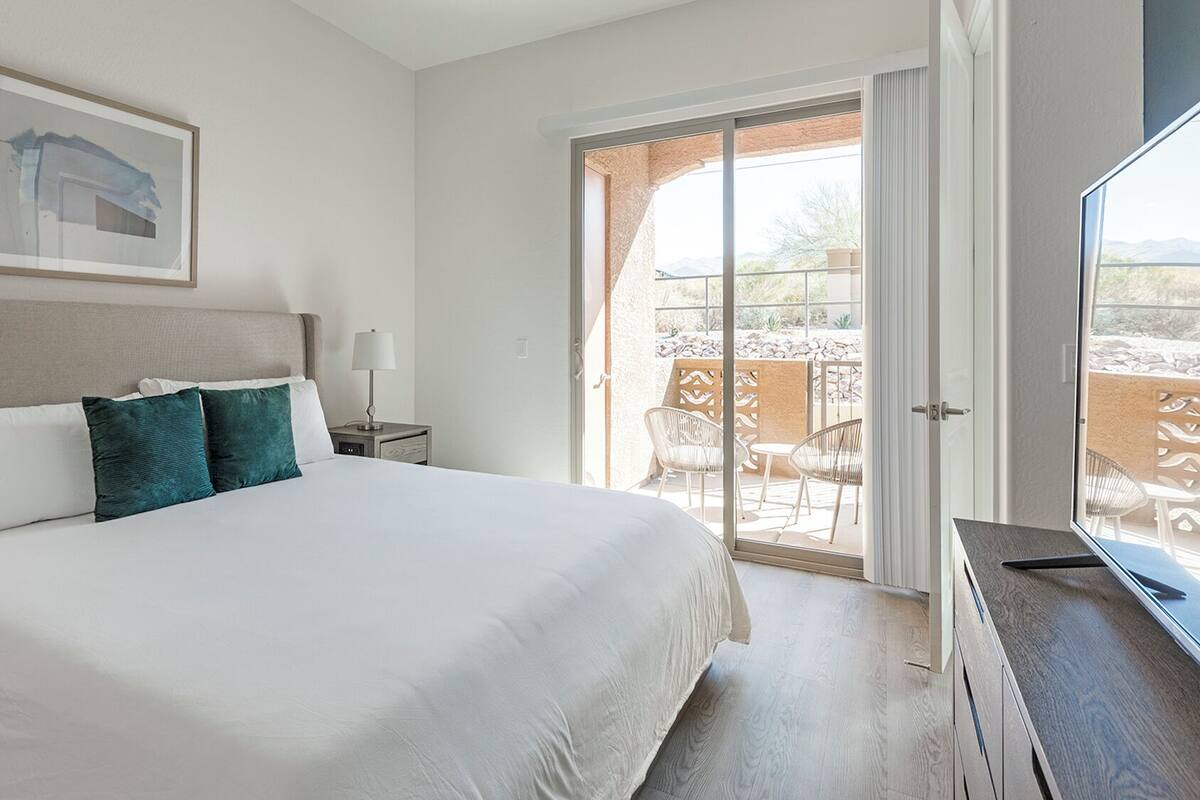 Une chambre d’hôtel avec un grand lit, une table de chevet, une lampe et une porte-fenêtre coulissante donnant sur un espace extérieur aménagé avec des chaises et une table.