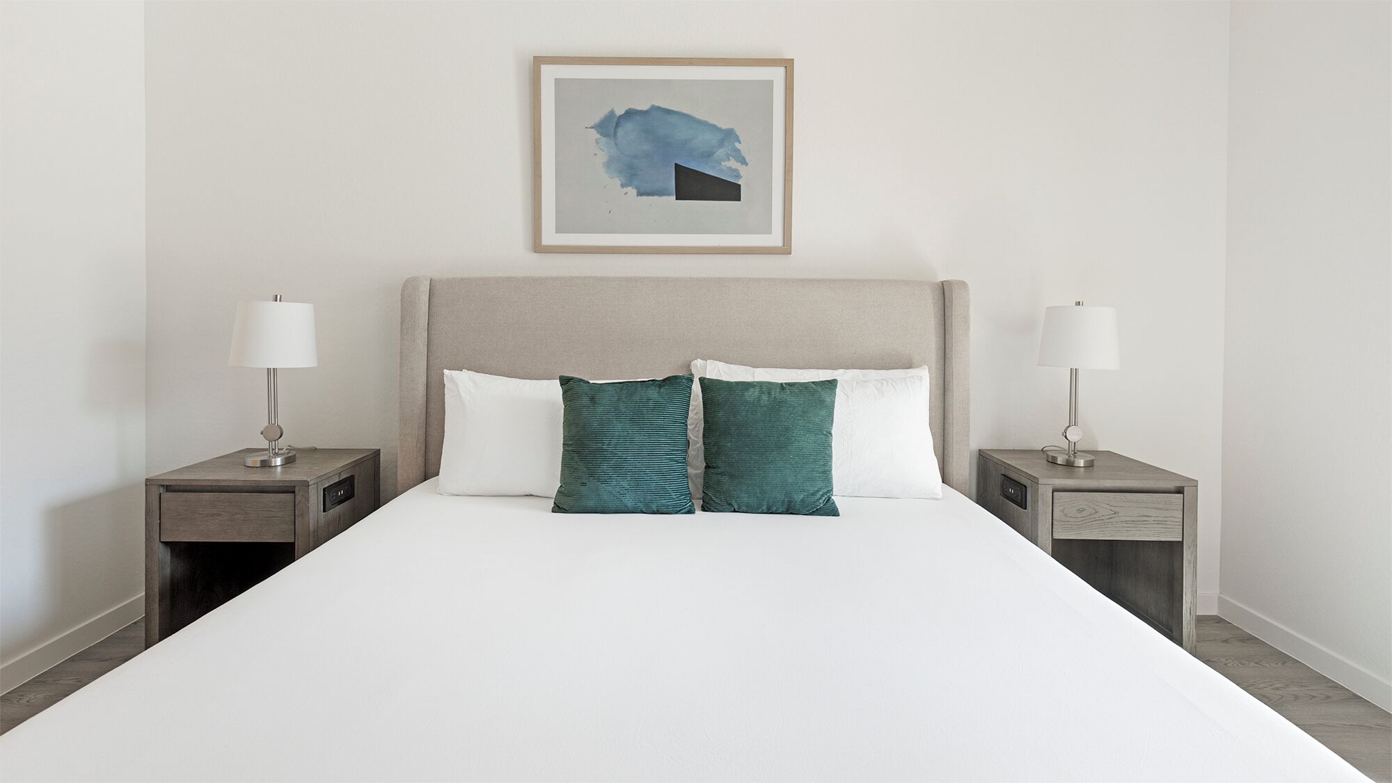 A neatly made bed with white linens, two green pillows, and a framed abstract painting above it.