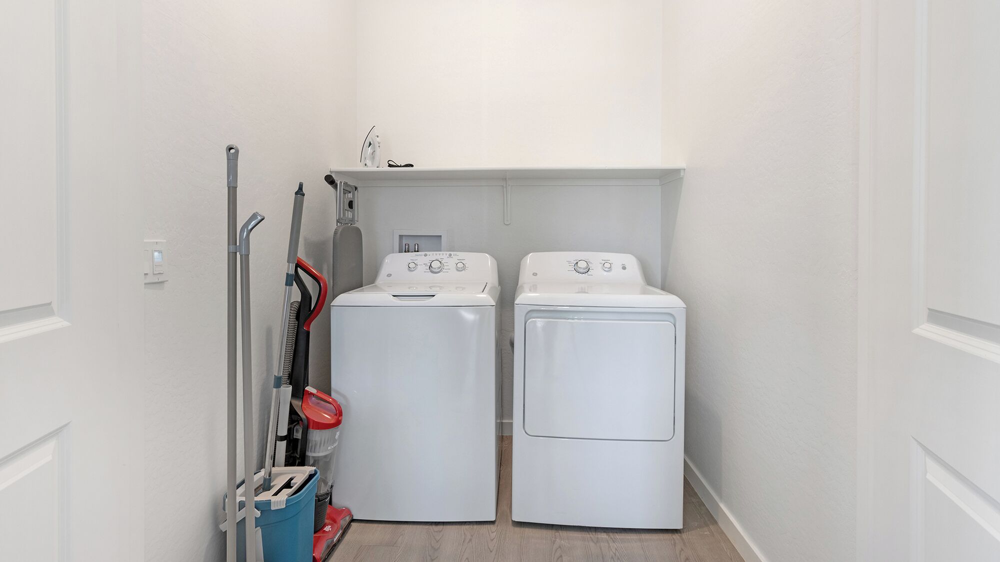 A laundry room with two white front-loading washing machines, a red and blue vacuum cleaner, and a mop.