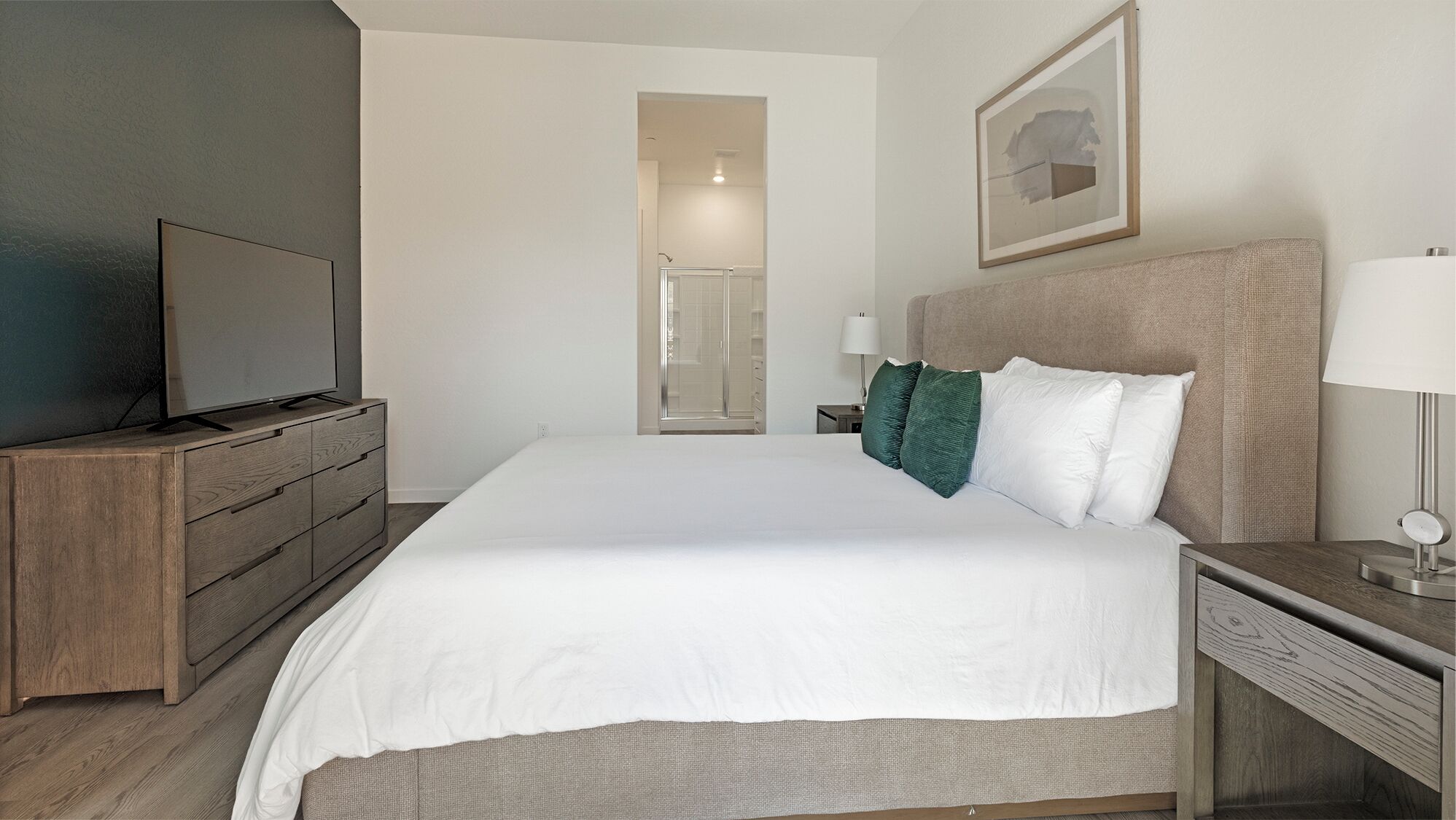 Une chambre d’hôtel avec un lit, un téléviseur posé sur une commode en bois, une table de chevet avec une lampe et un tableau encadré accroché au mur.