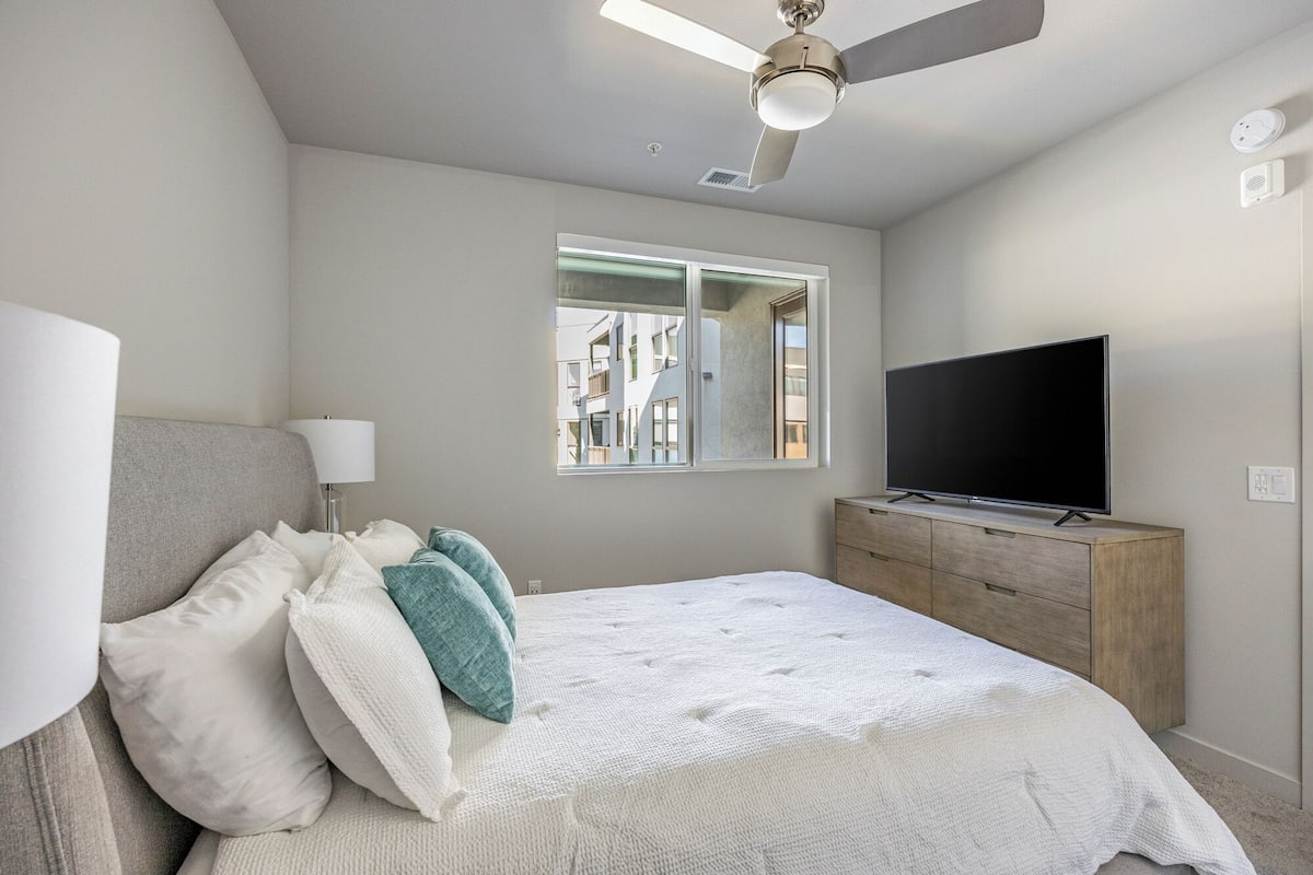 A bedroom with a bed, a TV on a wooden dresser, a ceiling fan, and a window with a view of buildings.