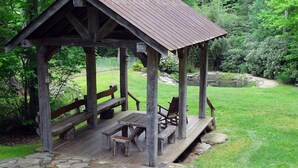 A rustic wooden gazebo with benches and picnic tables, surrounded by a stone-paved area and lush greenery.