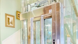 Un ascenseur moderne doté de portes en verre et d’une structure métallique, situé dans un immeuble qui possède un escalier et des tableaux encadrés accrochés au mur.